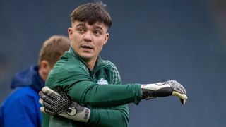 Ein Fußballspieler in einem grünen Trikot und Handschuhen steht draußen und schaut nach vorne. Hinter ihm ist ein anderer Spieler teilweise zu sehen.