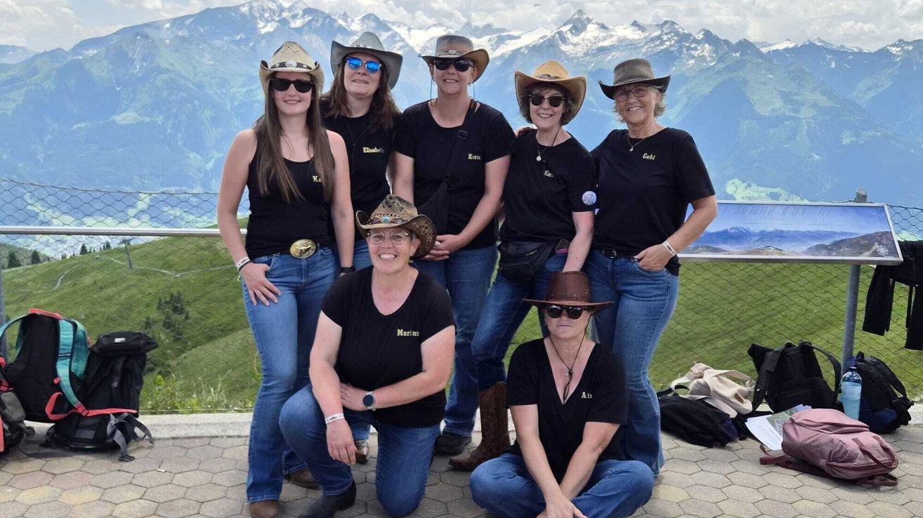 Eine Gruppe von Frauen in Cowboyhüten posiert für ein Foto vor einer Berglandschaft. Sie sind in schwarzen Shirts und Jeans gekleidet.