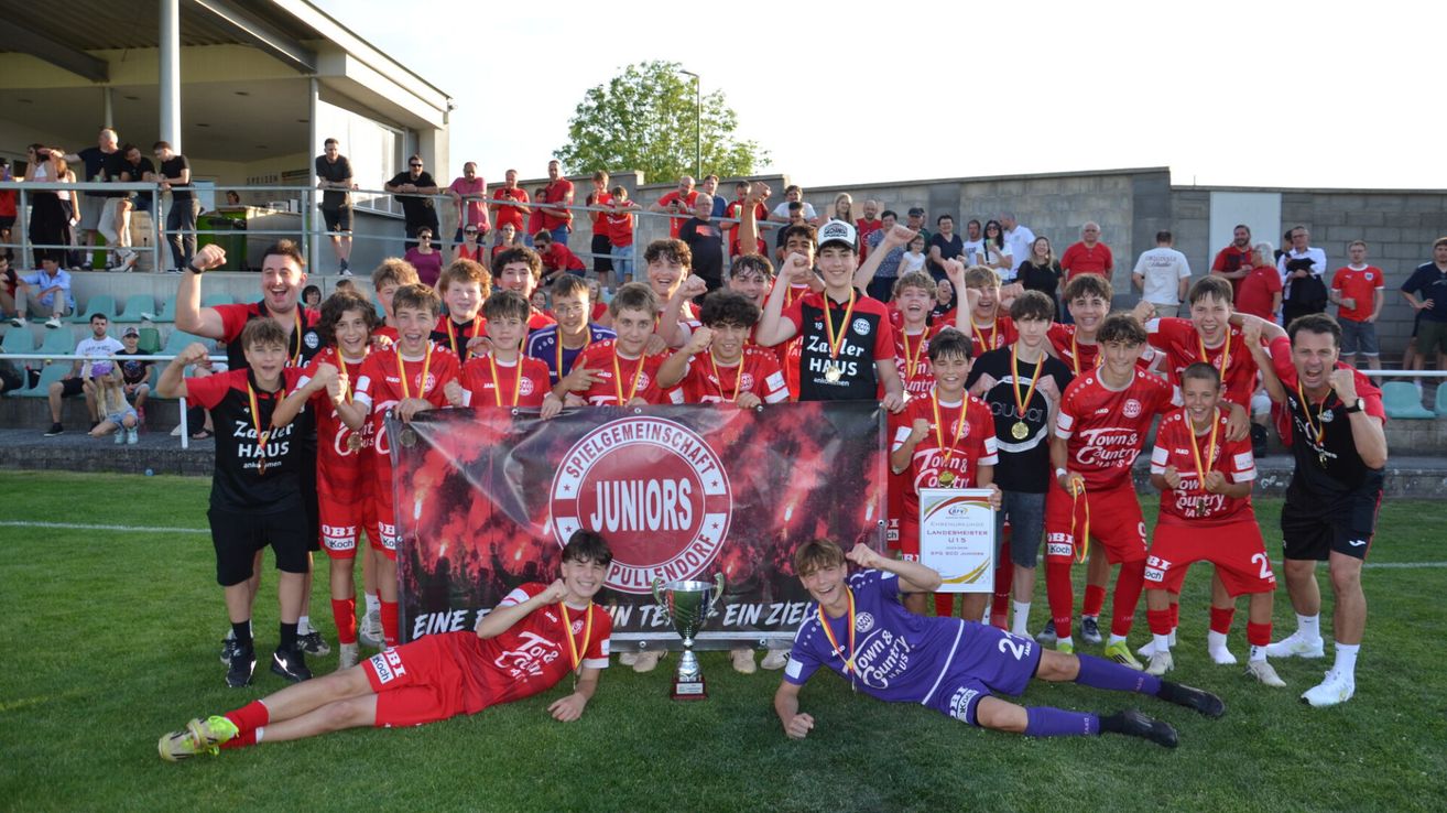 Eine Gruppe junger Fußballspieler in roten Trikots mit Medaillen posiert mit einem Banner, das 'Spielgemeinschaft Juniors Pullendorf' steht. Zwei Spieler in lila Uniformen liegen mit einem Pokal auf dem Boden. Eine Menge Menschen steht dahinter.
