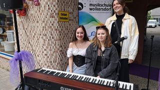 Drei junge Frauen stehen neben einem Klavier. Eine spielt es, während die anderen neben ihr stehen. Dahinter befindet sich eine Wand mit einem Schild, das 'Einfahrt freihalten!' sagt, und einem Banner für die 'Regionalmusikschule Wolkersdorf.'