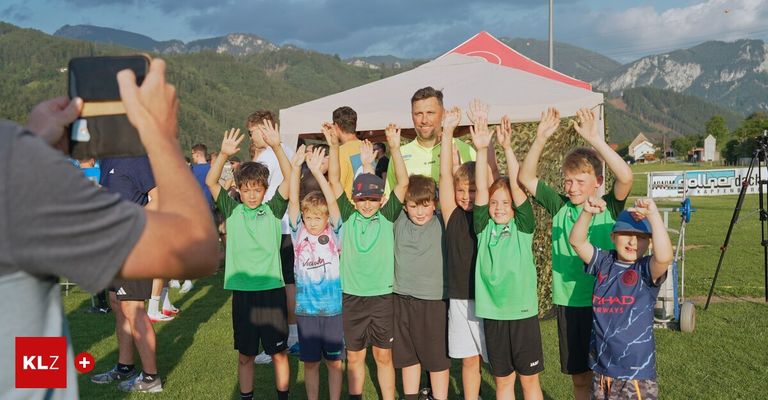 Eine Gruppe junger Jungen und Männer in grünen Trikots, einige mit Mützen, stehen unter einem Zelt mit erhobenen Händen. Sie scheinen bei einer Sportveranstaltung mit einem bergigen Hintergrund zu sein.