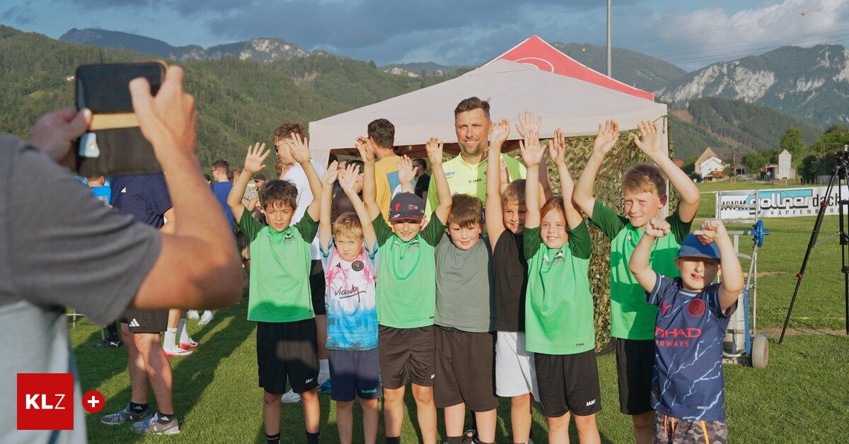 Eine Gruppe junger Jungen und Männer in grünen Trikots, einige mit Mützen, stehen unter einem Zelt mit erhobenen Händen. Sie scheinen bei einer Sportveranstaltung mit einem bergigen Hintergrund zu sein.