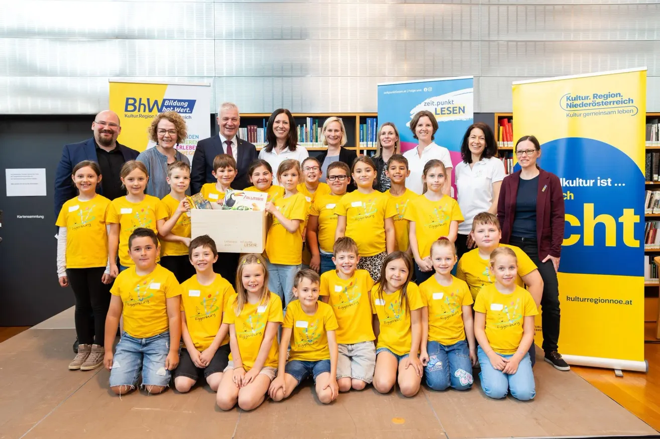 Eine Gruppe von Kindern in gelben T-Shirts posiert für ein Foto mit Erwachsenen, darunter ein Mann im Anzug und eine Frau in einem weißen Shirt. Sie befinden sich in einer Bibliothek mit Bücherregalen im Hintergrund.