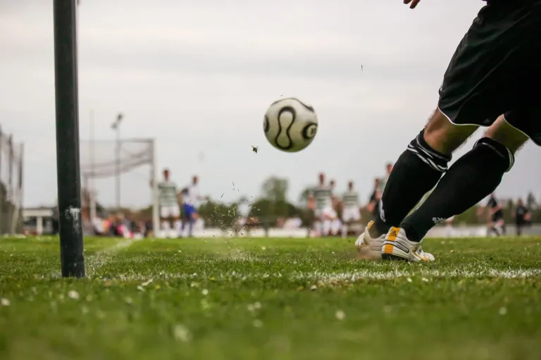 Ein Fußballspieler tritt einen Ball in der Luft. Der Ball ist leicht verschwommen. Die Füße des Spielers stehen auf dem Rasenfeld. Im Hintergrund stehen andere Spieler in der Nähe des Tores.