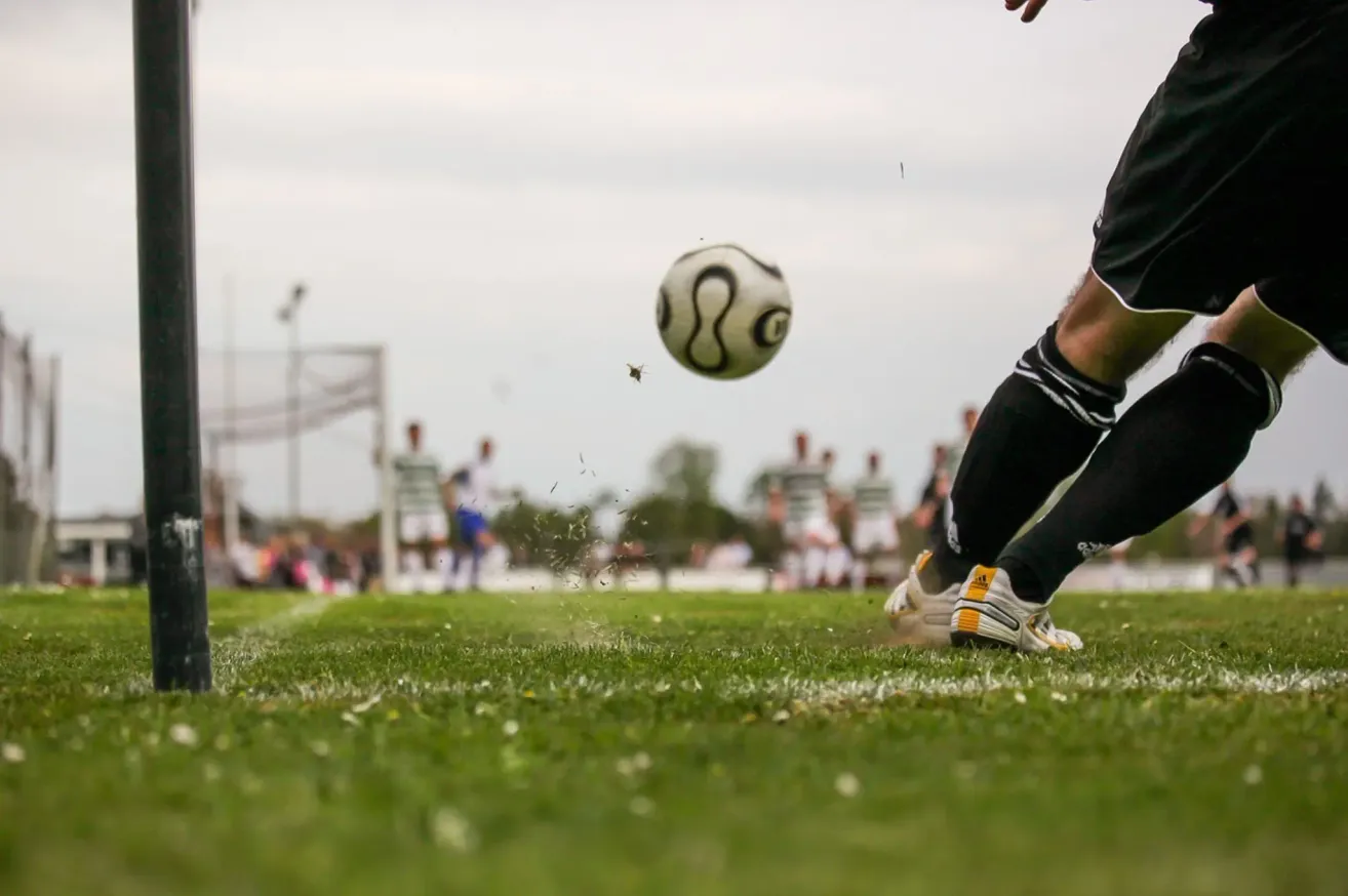 Ein Fußballspieler tritt einen Ball in der Luft. Der Ball ist leicht verschwommen. Die Füße des Spielers stehen auf dem Rasenfeld. Im Hintergrund stehen andere Spieler in der Nähe des Tores.