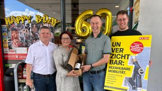 Drei Personen stehen vor einem Geschäft und feiern einen Geburtstag. Ein Mann in der Mitte hält eine Flasche Wein, während eine Frau links lächelt und ein Mann rechts ein Schild hält.