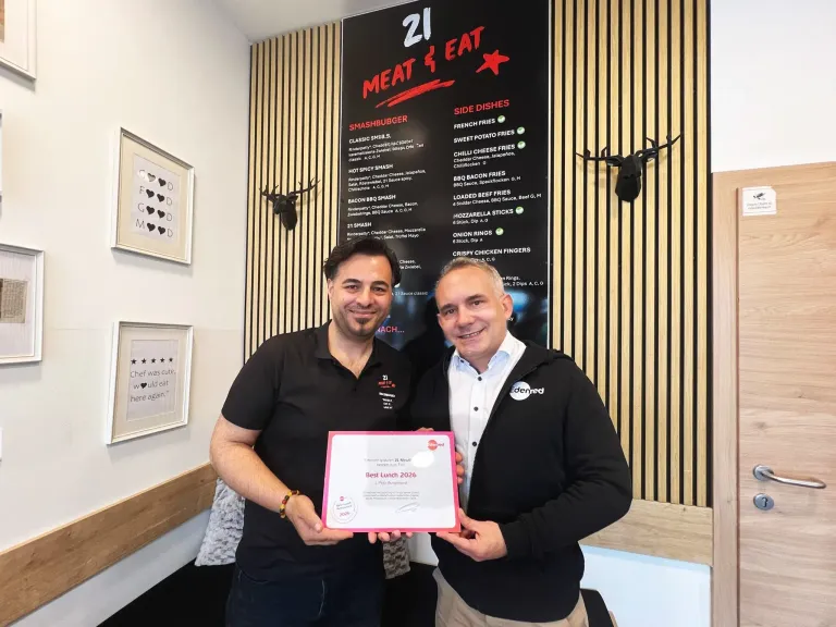 Zwei Männer halten ein Zertifikat mit 'Bestes Mittagessen 2026' in der Hand. Dahinter zeigt eine Wand eine Speisekarte mit verschiedenen Gerichten.