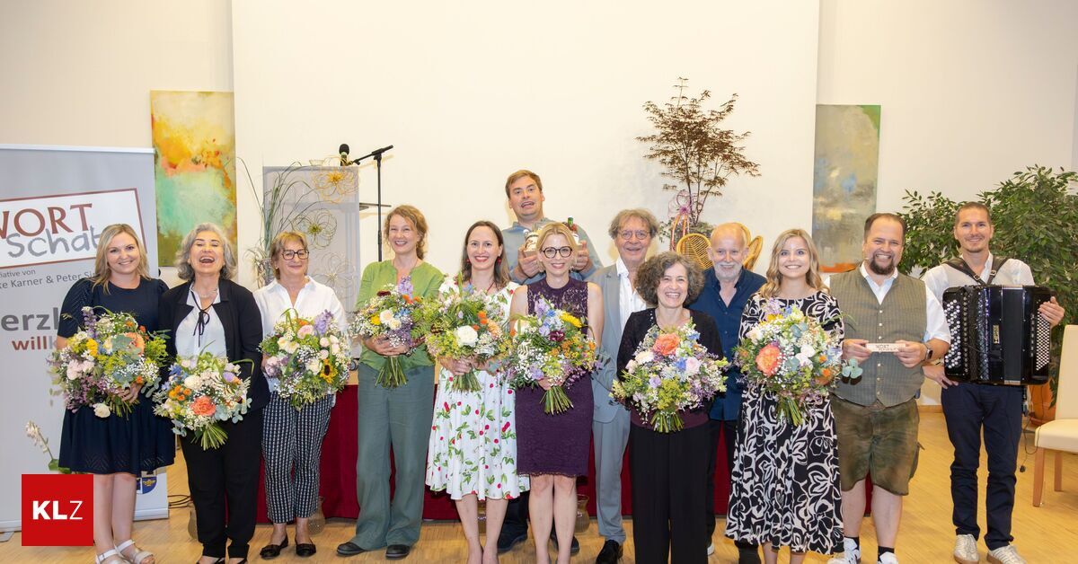 Eine Gruppe von Menschen, die lächeln und Blumensträuße halten, steht vor einer Bühne mit einem Mikrofon. Einige sind lässig gekleidet, während andere formelle Kleidung tragen. Hinter ihnen hängen Kunstwerke an der Wand.