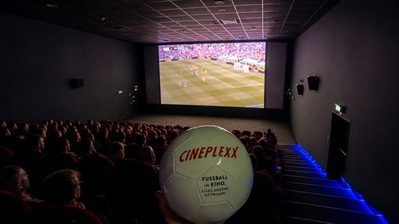 Eine Person hält einen Fußball mit der Aufschrift 'CINEPLEXX' vor eine Kinoleinwand, die ein Fußballspiel zeigt, in einem dunklen Kinosaal.