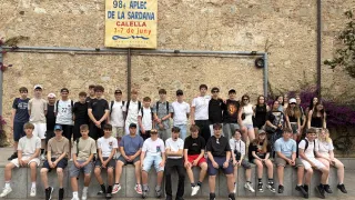 Eine Gruppe von Schülern der 98. APLC von La Sardana posiert für ein Foto. Sie sitzen auf einer Mauer mit einem Banner im Hintergrund.