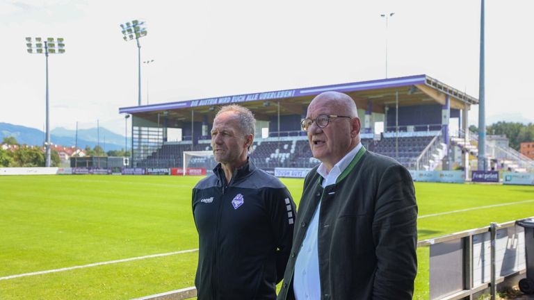 Zwei Männer stehen vor einem Stadion, einer in einer schwarzen Jacke, der andere in einer grünen Jacke mit weißem Innenteil. Sie stehen vor einem Fußballfeld mit grünem Rasen. Hinter ihnen befindet sich ein Stadion mit einer großen Bannerfahne.