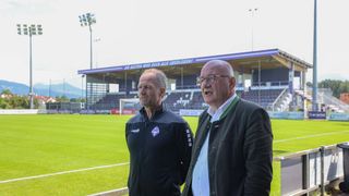 Zwei Männer stehen vor einem Stadion, einer in einer schwarzen Jacke, der andere in einer grünen Jacke mit weißem Innenteil. Sie stehen vor einem Fußballfeld mit grünem Rasen. Hinter ihnen befindet sich ein Stadion mit einer großen Bannerfahne.