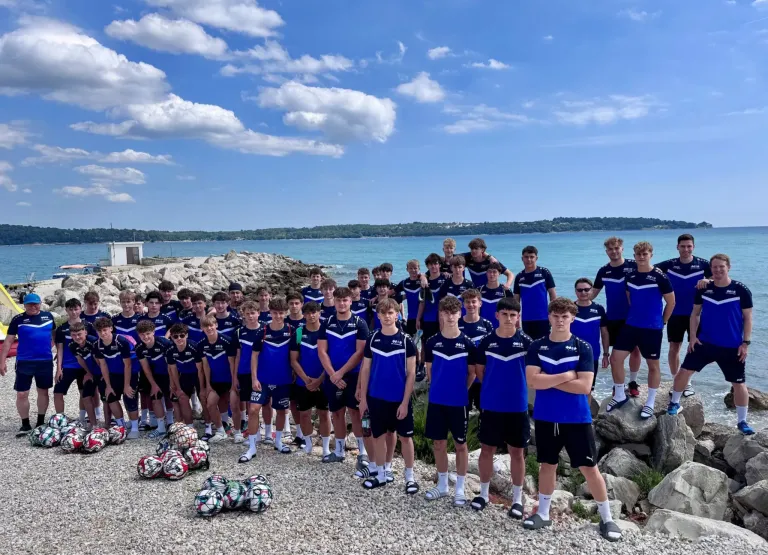 Eine Gruppe junger Männer in blauen Uniformen posiert für ein Foto am Meer. Einige stehen, andere sitzen auf Felsen. Dahinter befindet sich ein Gewässer mit Blick auf den Horizont.