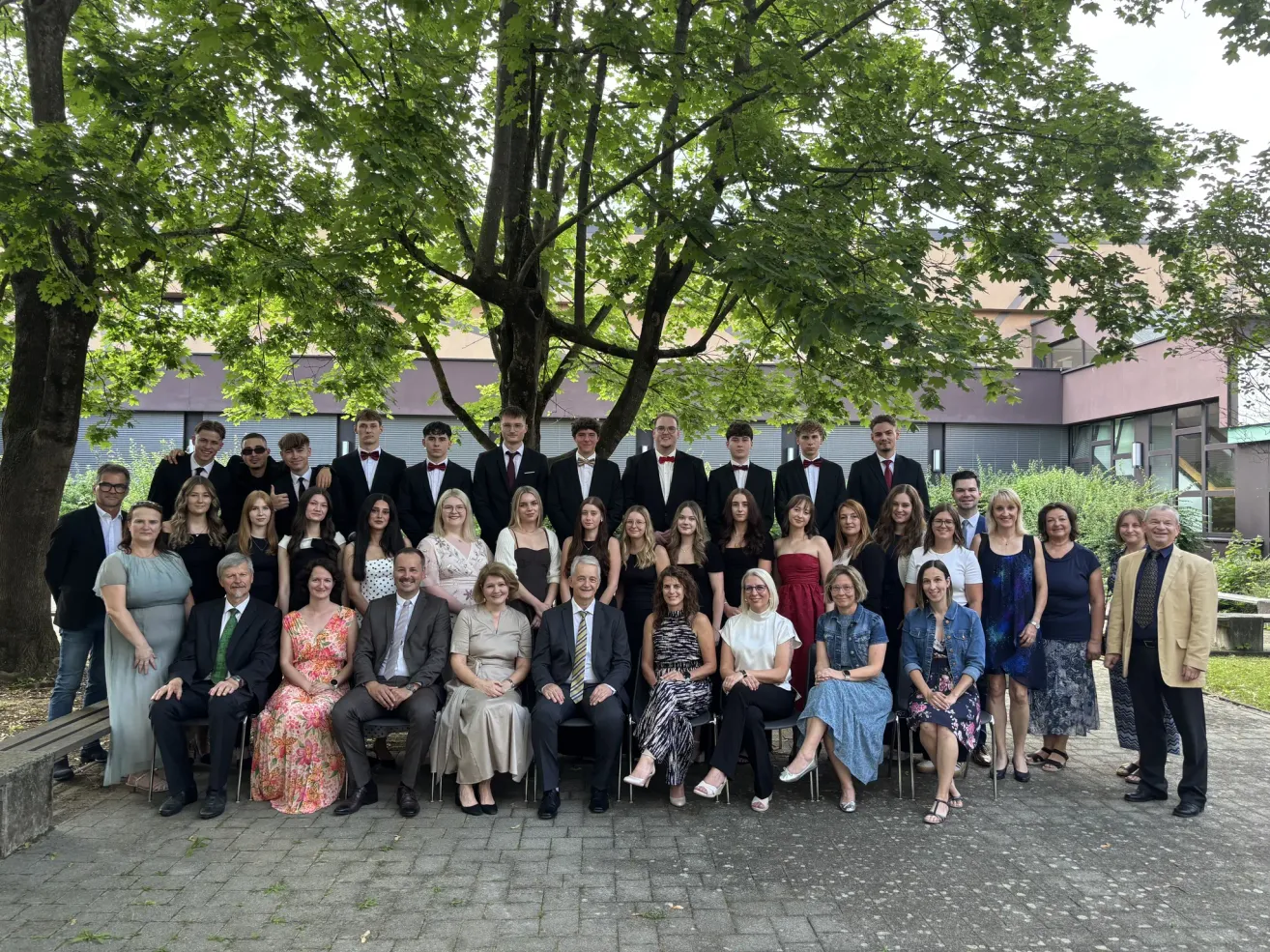 Eine Gruppe von Menschen, darunter Schüler und Lehrer, posiert für ein Foto unter einem Baum. Sie sind formell gekleidet, einige tragen Krawatten. Im Hintergrund befindet sich ein Gebäude.
