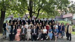 Eine Gruppe von Menschen, darunter Schüler und Lehrer, posiert für ein Foto unter einem Baum. Sie sind formell gekleidet, einige tragen Krawatten. Im Hintergrund befindet sich ein Gebäude.