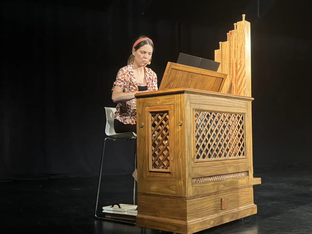 Eine Frau mit Stirnband spielt eine Holzorgel, während sie auf einer Bühne auf einem Stuhl sitzt. Die Orgel hat komplizierte Designs und einen schwarzen Laptop darauf.