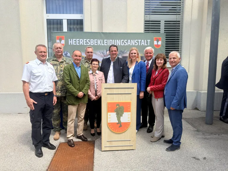 Gruppe von Menschen steht vor einem Gebäude mit einem Schild, auf dem Heeresbekleidungsanstalt steht. Vor ihnen steht ein Podest mit einem Schild.