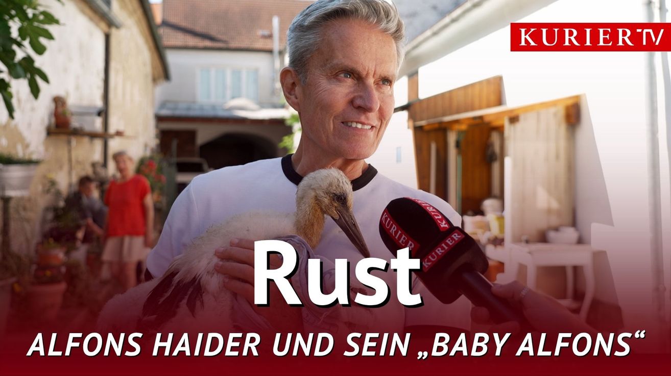 Ein Mann hält einen Storch vor ein Mikrofon. Er lächelt und hält den Vogel dicht an seiner Brust. Hinter ihm befindet sich ein Haus mit einem roten Dach. Die Wörter Rust Haider und sein Baby sind in weißen Buchstaben angezeigt.