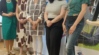 Fünf Frauen stehen eng nebeneinander vor einer Kulisse. Sie alle lächeln und posieren möglicherweise für ein Foto. Die Kulisse zeigt ein farbenfrohes Design mit Text und Bildern.