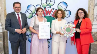 Vier Personen stehen zusammen vor einer Wandmalerei. Eine Frau hält ein gerahmtes Zertifikat. Eine andere Frau hält ein Schild mit der Aufschrift 'ZEICHENWEIN.' Der Mann trägt einen Anzug und eine Krawatte. Die vierte Frau trägt ein rotes Outfit.