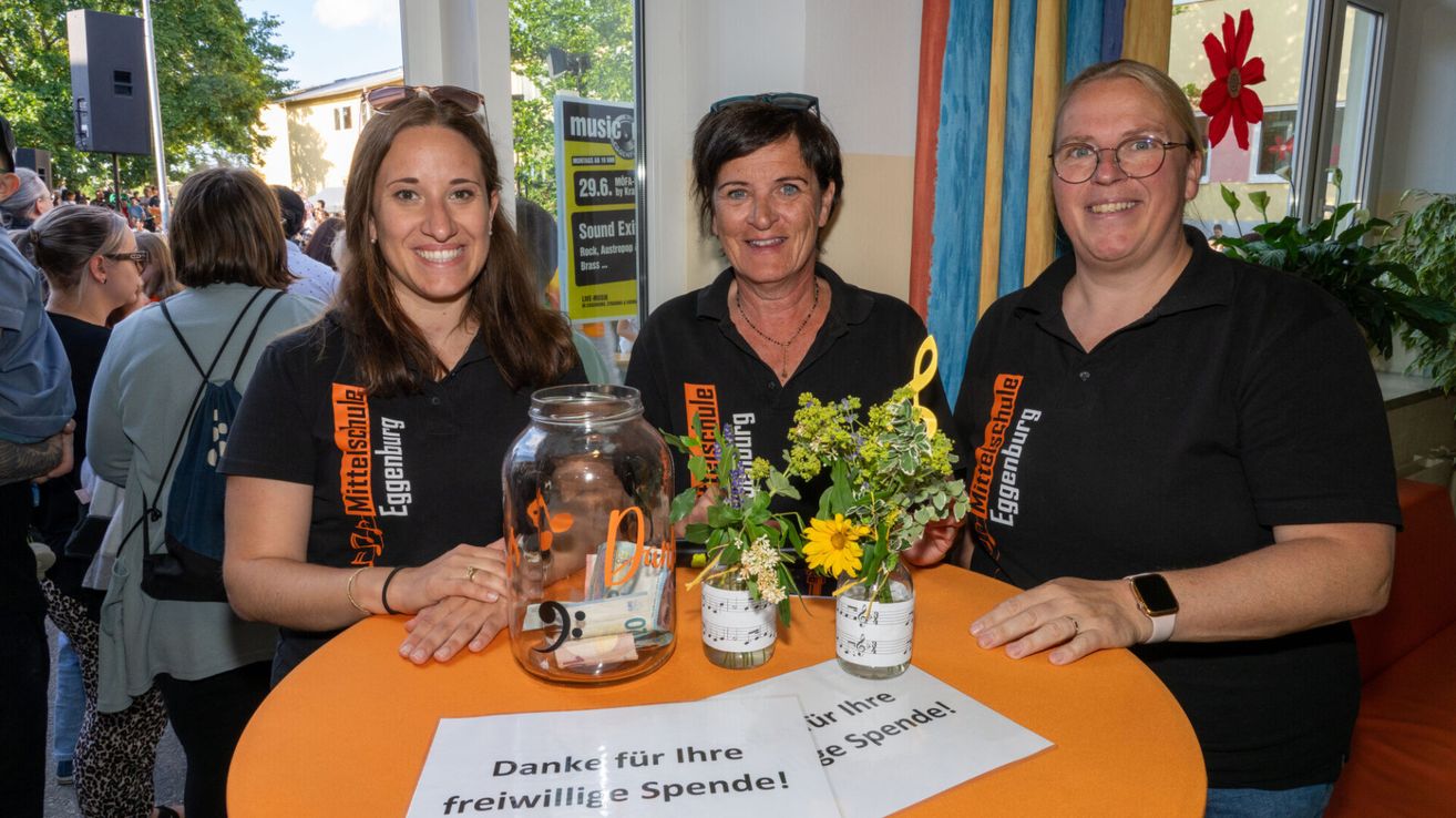 Drei Frauen lächeln für ein Foto und halten Geldgläser. Der Tisch hat ein Schild mit der Aufschrift 'Danke für Ihre freiwillige Spende!' Es gibt Blumen in den Gläsern.