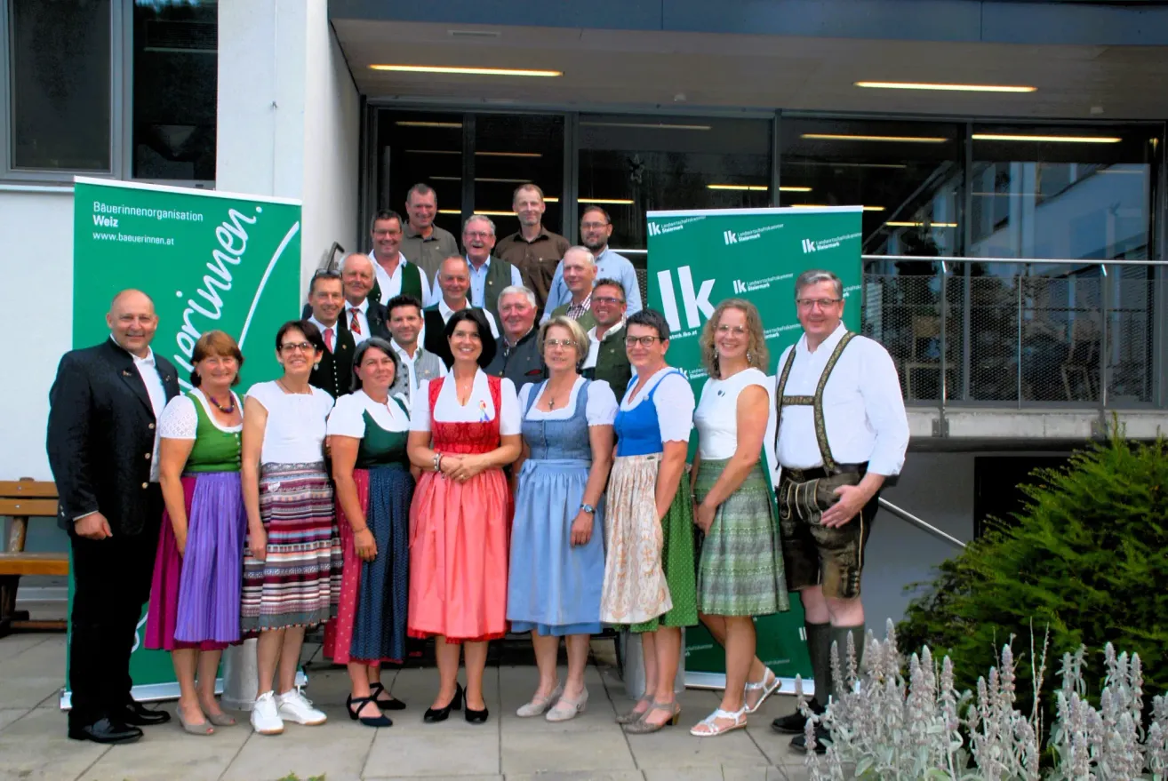 Eine Gruppe Erwachsener in traditioneller Kleidung posiert für ein Foto vor einem Gebäude mit großen Fenstern und grünen Bannern.