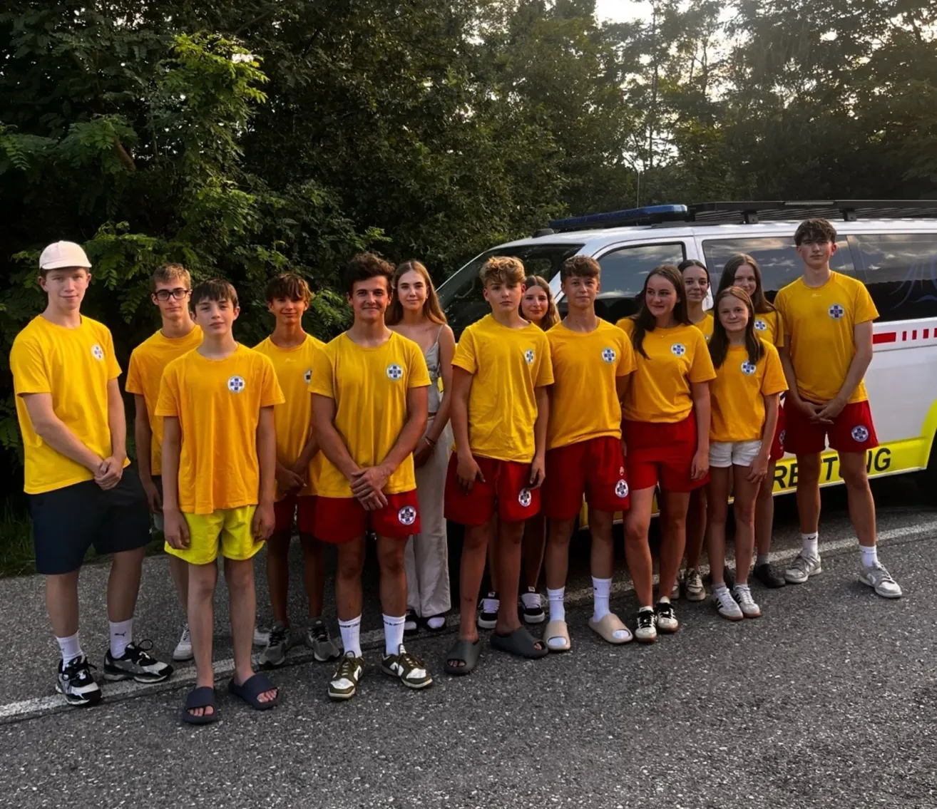 Eine Gruppe von Teenagern in gelben und roten Uniformen posiert für ein Foto vor einem weißen Van. Bäume sind im Hintergrund zu sehen.