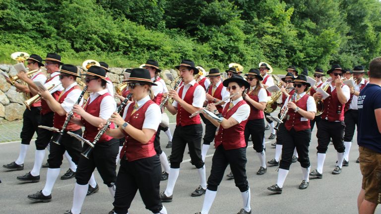 Eine Blaskapelle in roten Westen und schwarzen Hüten spielt Instrumente und zieht in einer Parade auf einer Straße, mit Grün im Hintergrund.
