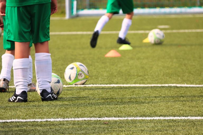 Zwei Fußballspieler in grünen Trikots stehen in der Nähe eines Torpfostens. Zwei Fußballbälle liegen auf dem Boden mit Kegeln in verschiedenen Farben.