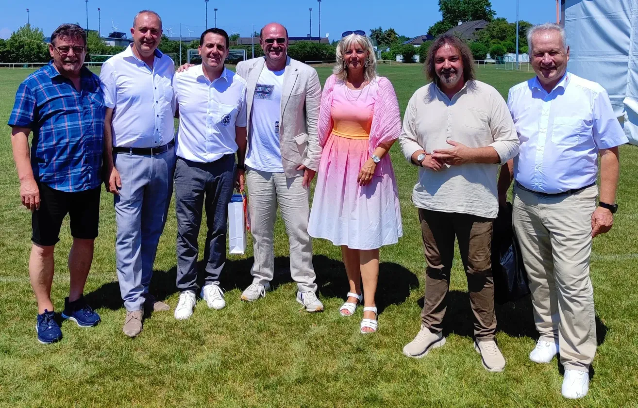 Fünf Personen stehen in einer Reihe auf einem Feld und lächeln für die Kamera. Die Frau trägt ein rosa Kleid und die Männer leichte Kleidung. Sie scheinen sich bei einer Outdoor-Veranstaltung zu befinden.