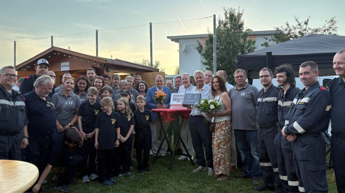 Eine Gruppe von Menschen steht in einem Park mit einem Tisch, Blumen und einer Plakette. Einige tragen Brillen und Uniformen.