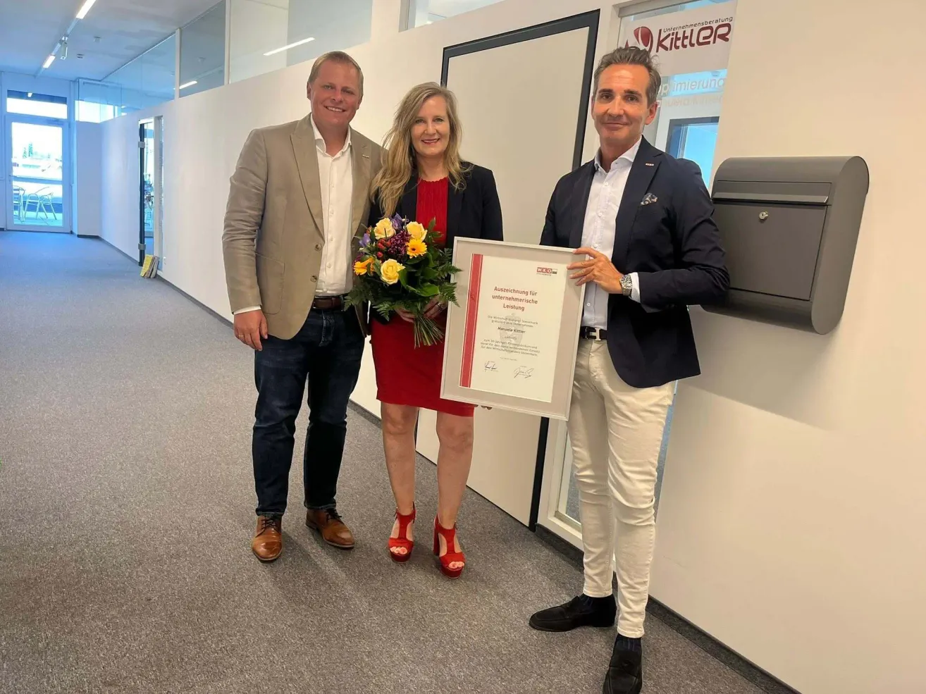Drei Personen posieren für ein Foto mit einem Zertifikat und Blumen. Die Frau trägt ein rotes Kleid, und der Mann neben ihr trägt einen beigefarbenen Anzug.