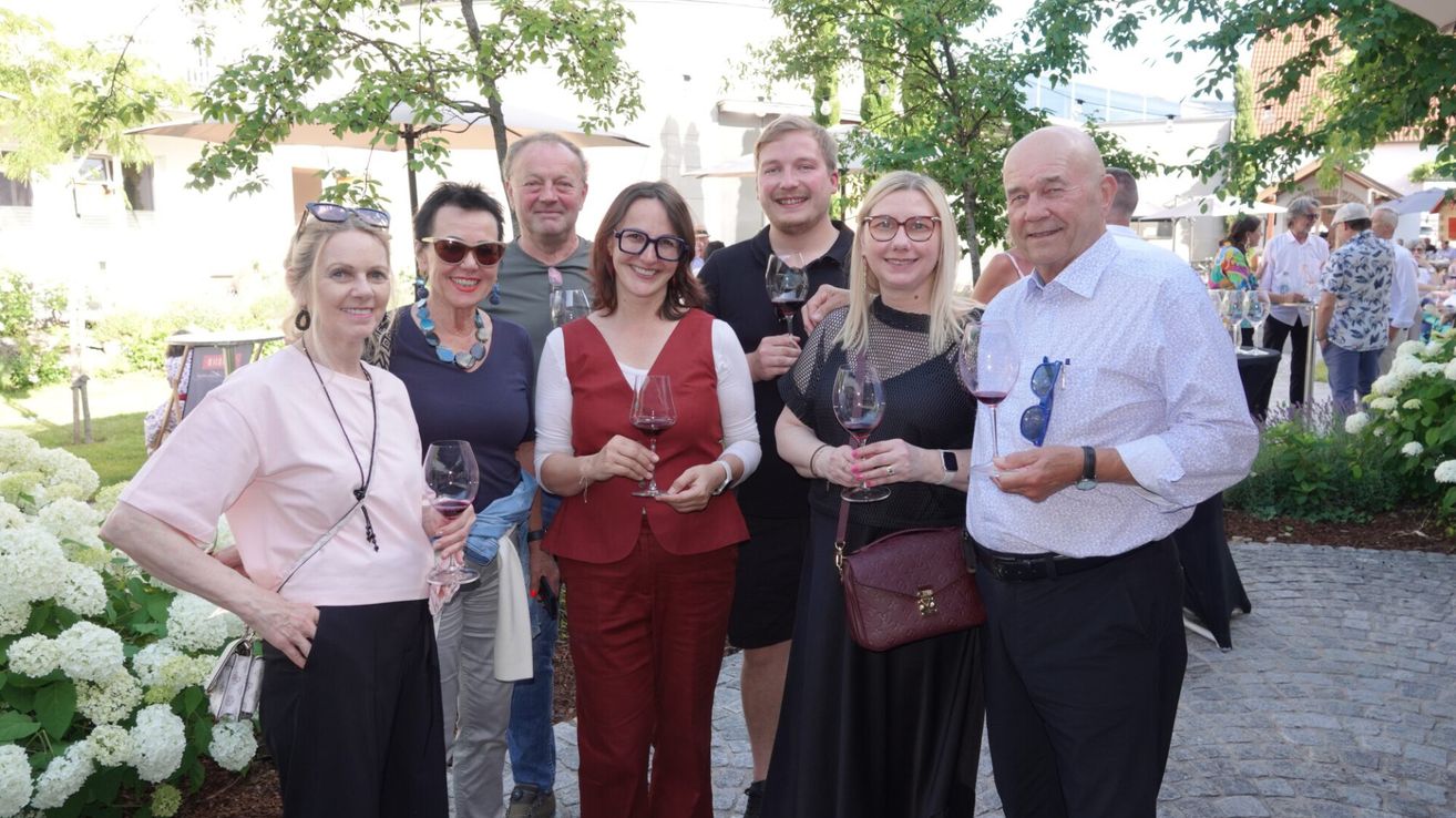 Eine Gruppe von Erwachsenen, sowohl Männer als auch Frauen, stehen zusammen. Sie halten Weingläser in den Händen, lächeln und scheinen für ein Foto zu posieren.