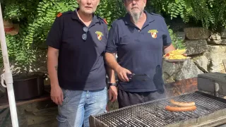 Zwei Männer in Uniformen stehen in der Nähe eines Grills im Freien, einer hält einen Teller mit Essen. Sie befinden sich in der Nähe einer Steinmauer und eines Baumes.