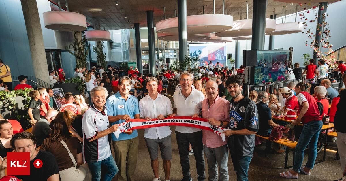 Fünf Männer stehen vor einer Menge und halten ein rotes Banner mit weißen Text. Sie lächeln alle und scheinen zu feiern. Der Mann ganz links trägt ein Fußballtrikot.
