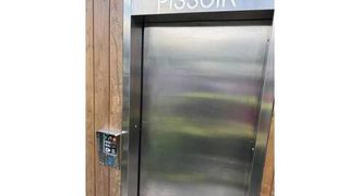 Ein Aufzug mit rostfreiem Edelstahlgehäuse und einer Holzwand. Oben steht 'PISSOIR' und links befindet sich ein Bedienfeld.