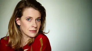 Eine Frau mit braunen Haaren und blauen Augen posiert für ein Foto. Sie trägt ein rotes Shirt mit goldenen Streifen und schaut nach vorne. Sie scheint in einem Studio zu sein.
