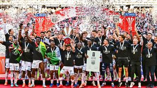 Fußballspieler in schwarzen Trikots feiern ihren Sieg. Sie halten einen Pokal, Medaillen und ein Zertifikat auf einer Bühne. Konfetti fällt von oben.
