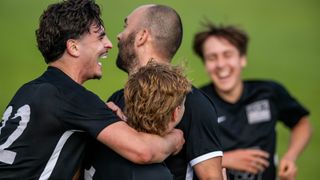 Vier junge Männer in schwarzen Trikots lachen und umarmen sich auf einem grünen Feld.