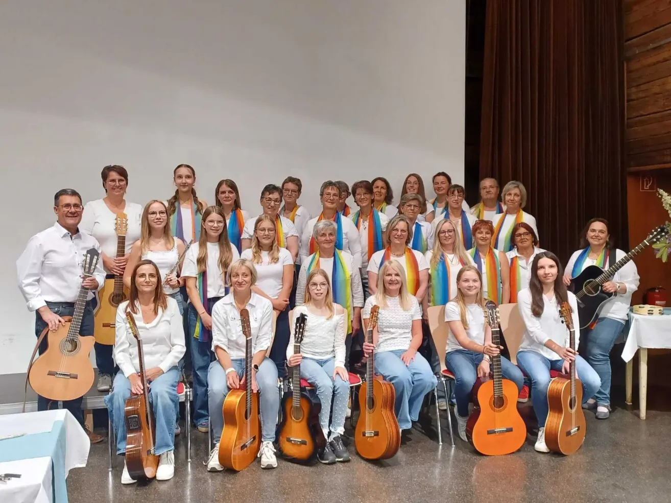 Eine Gruppe von Frauen, die weiße Hemden und Jeans tragen, sitzen mit ihren Gitarren auf Stühlen und posieren für ein Foto. Sie alle lächeln und tragen Regenbogen-farbene Schals.