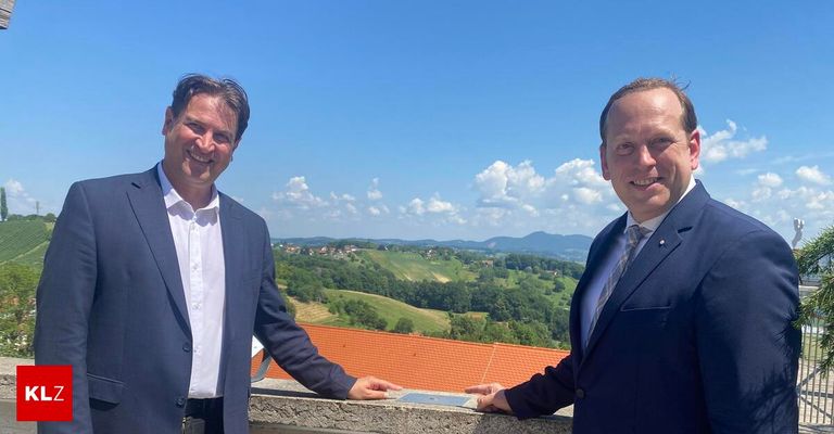 Zwei Männer in Anzügen stehen auf einem Balkon mit Blick auf eine bergige Landschaft.