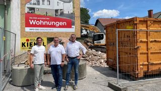 Drei Männer stehen in der Nähe einer Baustelle. Hinter ihnen ist ein Schild, das neue Wohnungen bewirbt. Ein Baufahrzeug ist im Hintergrund zu sehen.