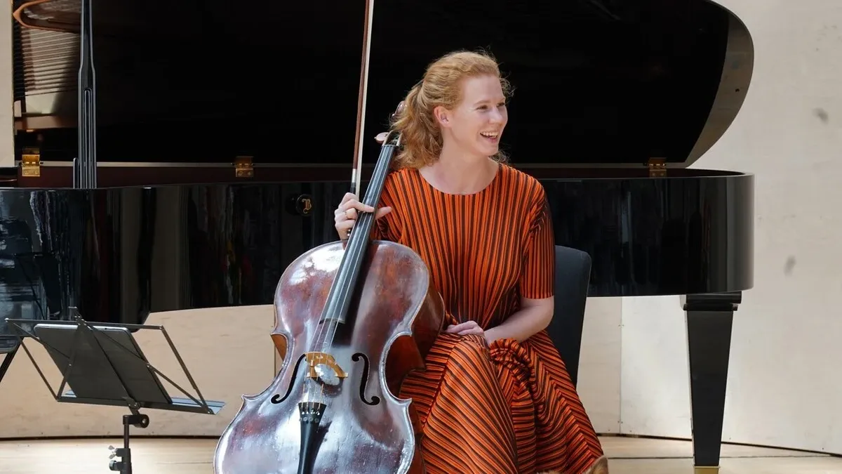 Eine Frau in einem orangefarbenen gestreiften Kleid sitzt auf einem Stuhl, hält ein Cello und lächelt. Der Hintergrund ist verschwommen und zeigt ein Klavier und eine schwarze Wand.
