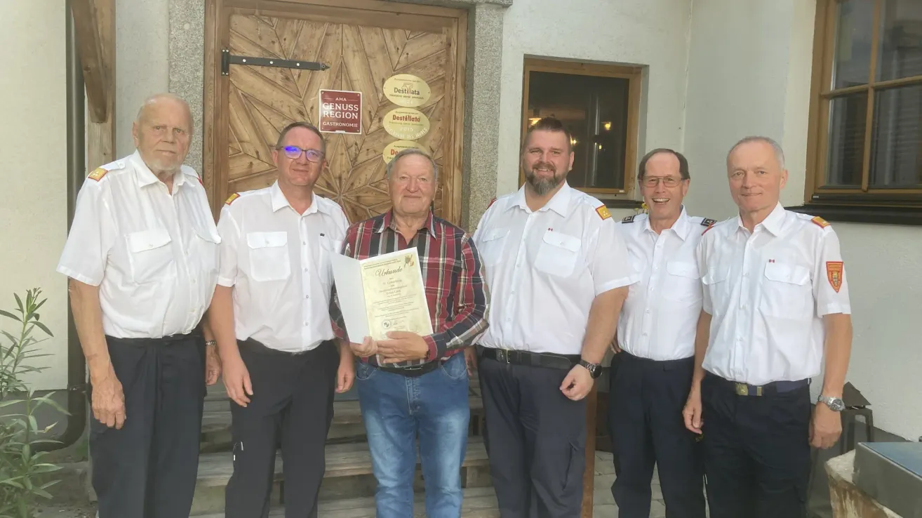 Fünf Männer stehen draußen, vier in Uniform und einer hält eine Urkunde. Sie stehen vor einer Holztür mit mehreren Schildern.