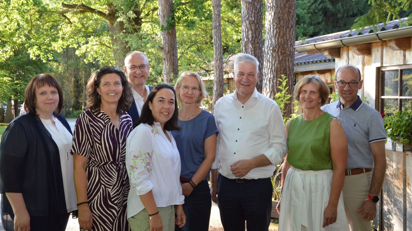 Eine Gruppe von sechs Personen, darunter drei Frauen und drei Männer, posiert für ein Foto in einem Park mit hohen Bäumen im Hintergrund.