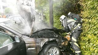 Feuerwehrleute arbeiten an einem verbrannten Auto, einer von ihnen untersucht den Motor, während der andere hinter ihm steht, beide in voller Feuerwehrausrüstung.