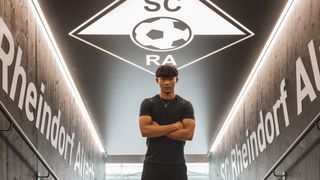 Ein Mann steht selbstbewusst in einer Sporteinrichtung mit dem SC Rheindorf Altach Logo über ihm, trägt ein schwarzes T-Shirt und eine Halskette.