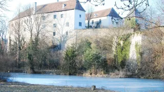 Ein großes weißes Gebäude steht auf einem Hügel, umgeben von laublosen Bäumen und einem gefrorenen See. Das Gebäude hat viele Fenster und ein braunes Dach.