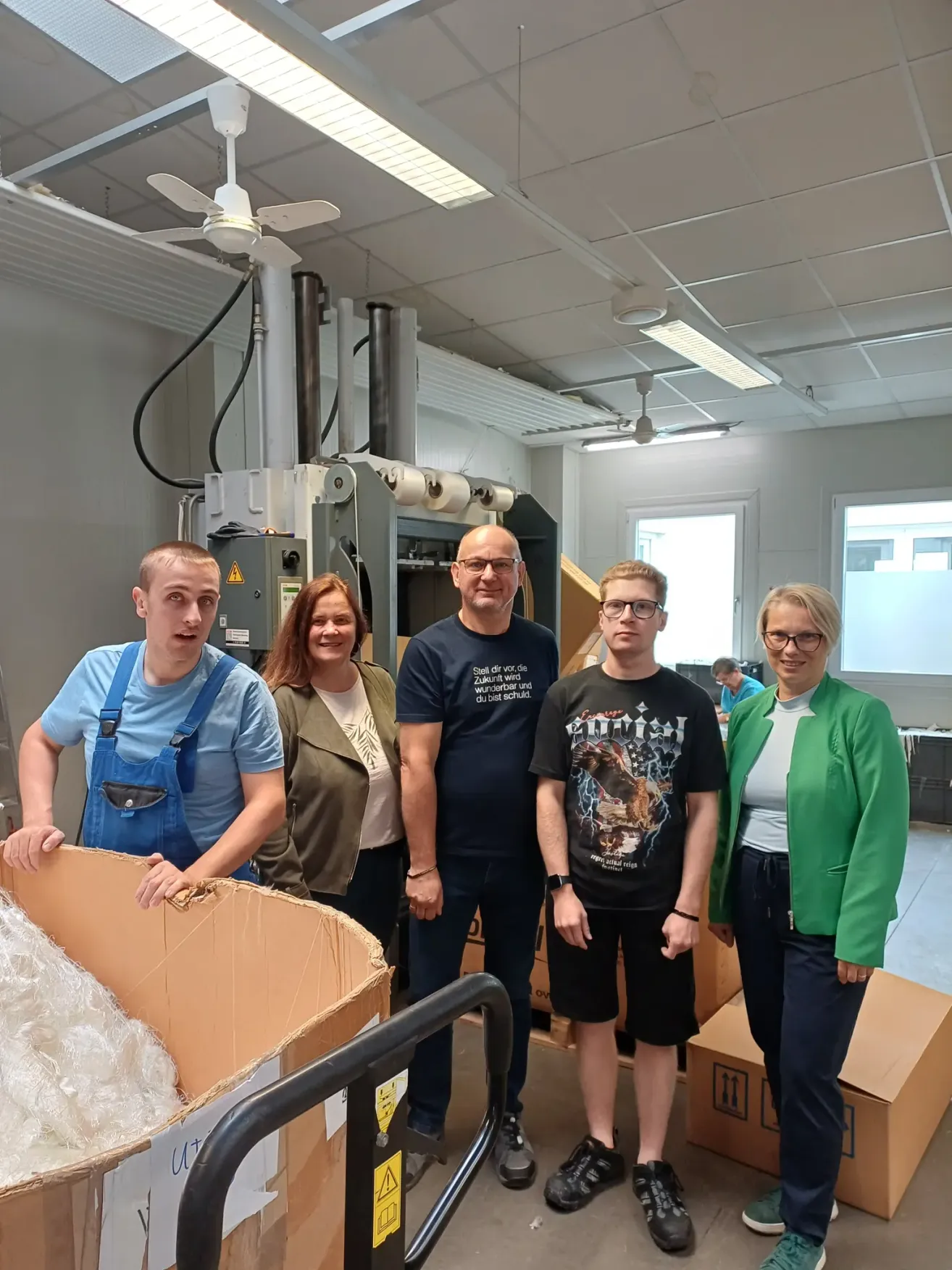 Fünf Personen stehen gemeinsam in einem Raum. Ein Mann auf der linken Seite hält eine Pappschachtel, während ein anderer Mann auf der rechten Seite einen Trolley hält. Hinter ihnen steht eine große Maschine mit Kabeln und einem Fenster.
