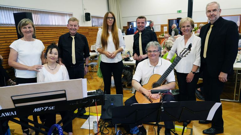 Eine Gruppe von Menschen steht lächelnd in einem Raum, für ein Foto. Musikinstrumente und Zubehör umgeben sie.